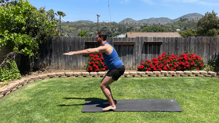 Man in a backyard on a yoga mat practicing Chair Pose with arms extended straight in front