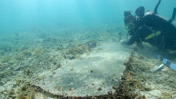 PHOTO: A scuba diver with measuring tools hovers alongside an underwater headstone surrounded by sand and short vegetation. Some letters and numbers can be made out in the inscription. (NPS Photo by C. Sproul)