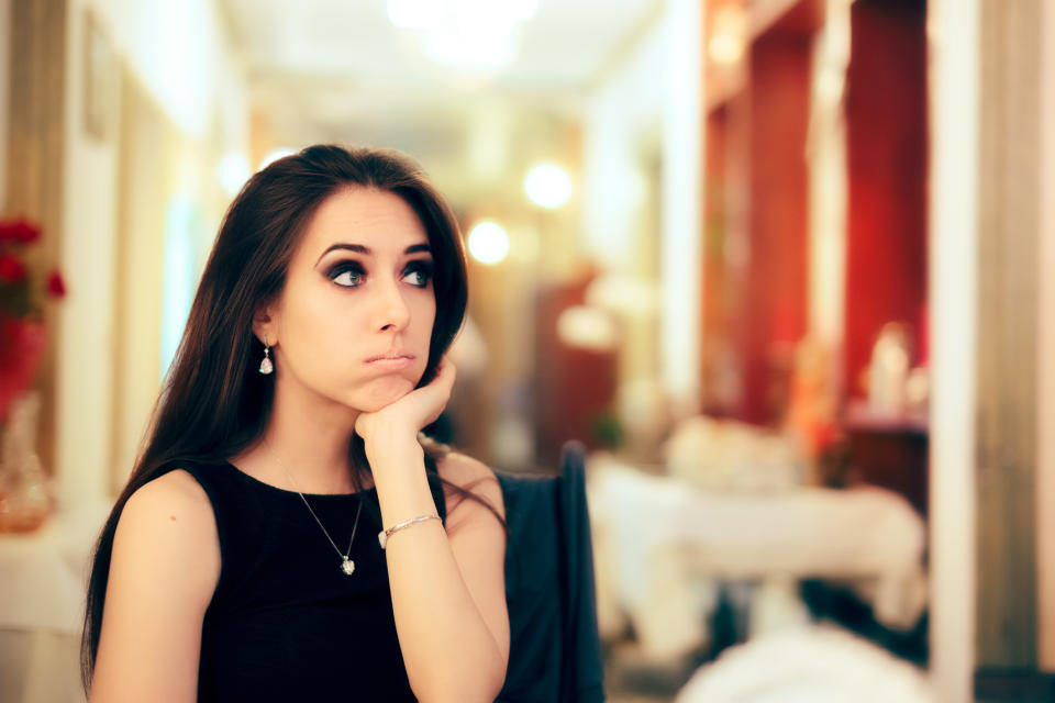 A woman with long hair wearing a sleeveless top and jewelry rests her chin on her hand, looking thoughtful in an elegant indoor setting