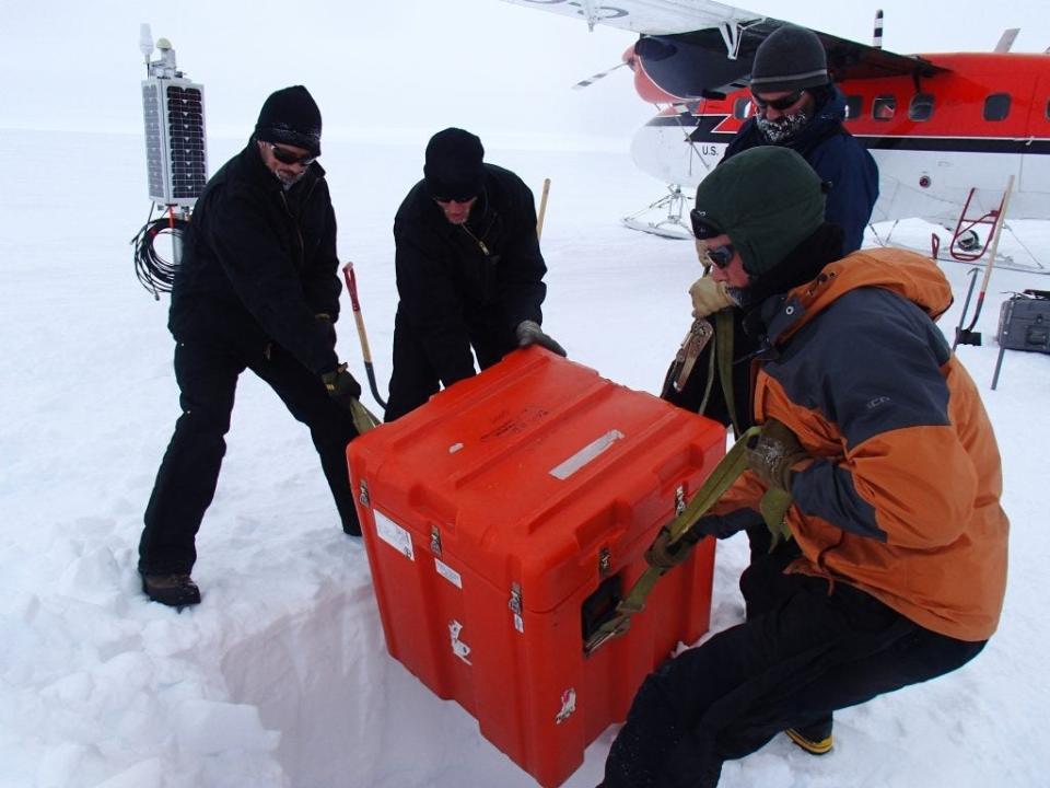 Researchers are shown wearing heavy snow gear in an icy environment in Antartica. A plane is seen in the background, fitting with landing gear for snowy terrain. The reserachers are seen lowering a heavy orange box — containing seismic detectors — into a hole in the snow.