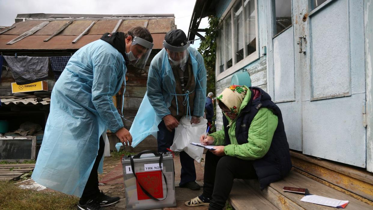 Zwei Wahlhelfer stehen neben einer Frau, die auf den Stufen vor einem Haus sitzt und Dokumente ausfüllt.