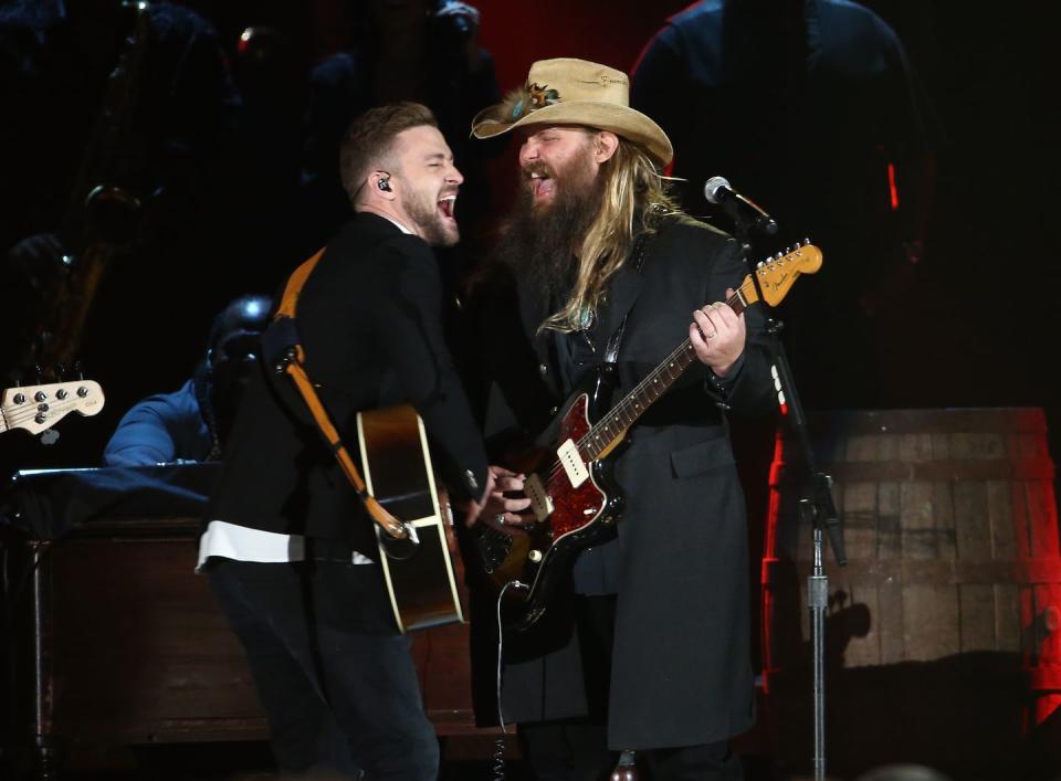 justin timberlake and chris stapleton stand and face each other on a stage, they sing toward a microphone on a stand and each play guitar
