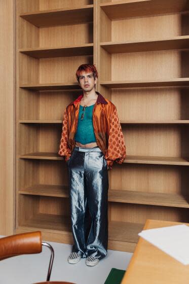 Julio Torres, in shiny silver pants, green tank and orange shirt, stands with hands in pockets before empty shelves