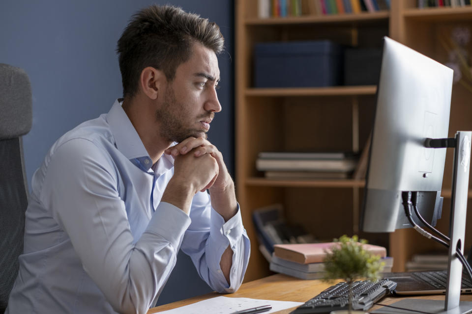 A man in a blue shirt sits at a desk, looking intently at a computer screen with his hands clasped under his chin. There are bookshelves in the background