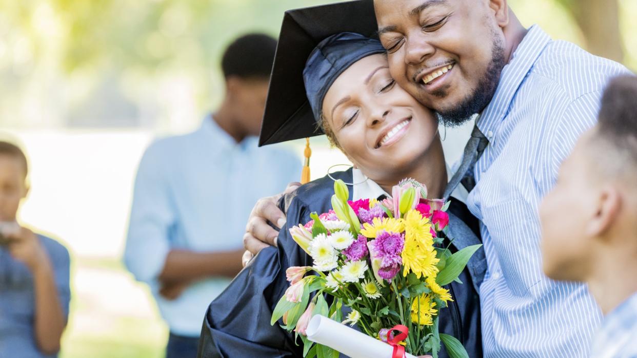  Man gives woman a bouquet of flowers on her graduation day. He is hugging her, while she is holding the flowers and a diploma. 