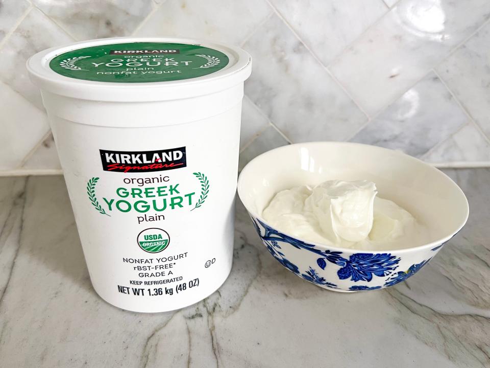 A large white container of Kirkland Signature organic plain Greek yogurt with a green lid. The container sits on a gray counter next to a blue-and-white bowl filled with white Greek yogurt