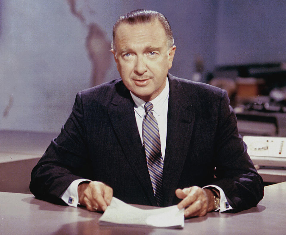 Walter Cronkite in a dark suit and striped tie, seated at a news desk, holding papers