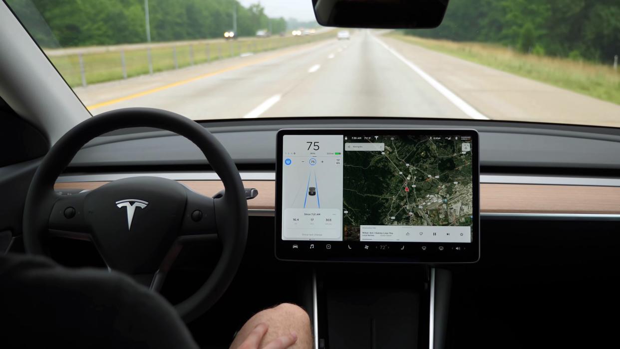 Missouri, United States - February 21, 2022: Close-up of a Tesla Model Y, driving on autopilot, down a highway straightaway surrounded by a forest, and a man sitting with his hands on his legs.