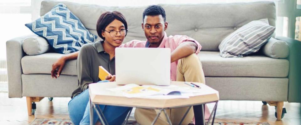 Dark skinned couple in love looking at mortgage rates on a laptop computer while sitting on their living room floor.