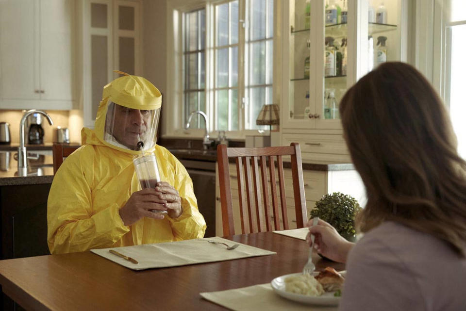 Shalhoub as Monk sits at a kitchen table in a yellow hazmat suit, sipping an iced coffee from a tumbler with a straw in a hole through his suit mask. (Courtesy of Peacock)