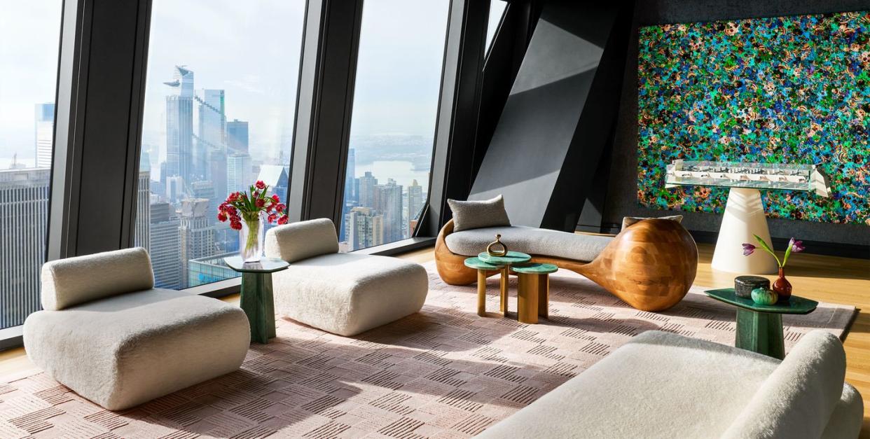 a penthouse living room has floor to ceiling windows, an armless sofa and chairs in an off white textured fabric, geometric rug, a curved wood chaise with fabric cushion, foosball table, large colorful painting