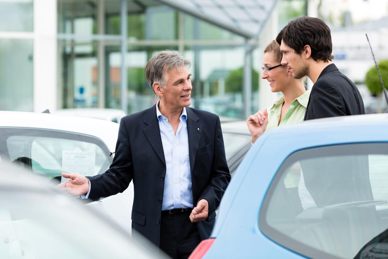 Mature male car dealer showing young couple cars in parking lot of car dealership with car dealership building in the background