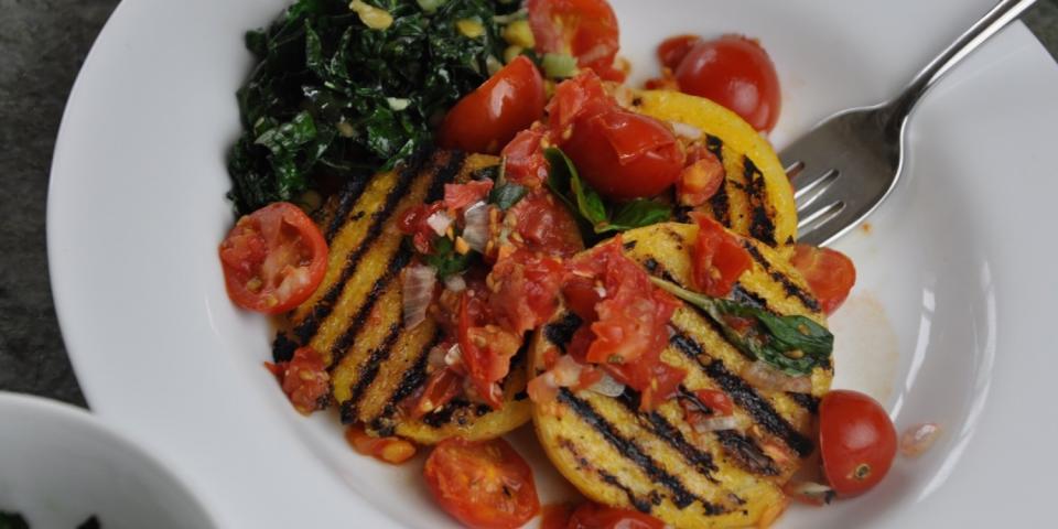 A plate of grilled polenta topped with cherry tomatoes, greens, and herbs, with a fork on the side