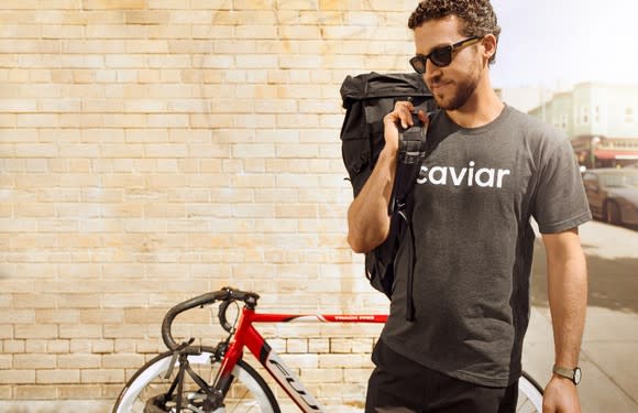 A Caviar employee carrying food in a bag over his right shoulder with a bike leaning on the wall behind him.