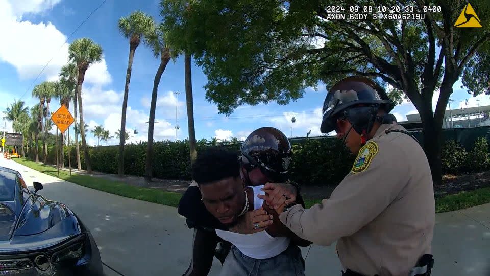 The officer puts his right arm around Hill and brings him down to sit on the sidewalk. - Miami Dade Police Department