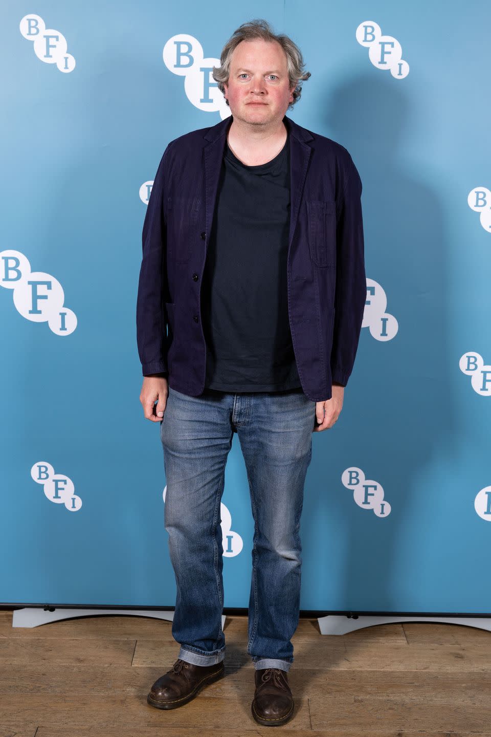 miles jupp wearing a black shirt and blazer with jeans, standing in front of a blue background with the bfi logo repeating over it