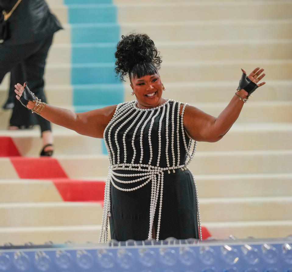 Lizzo on a staircase, wearing a black dress adorned with pearl strings and fingerless gloves, smiling and waving her hands up