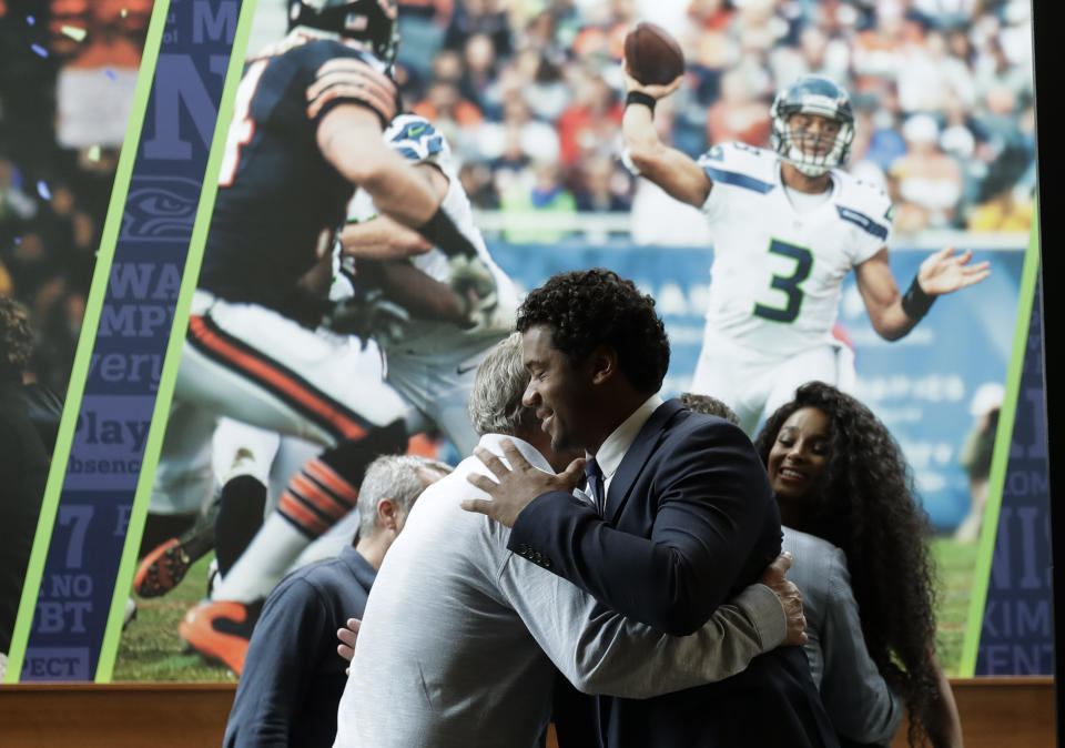 Seattle Seahawks quarterback Russell Wilson, center, hugs head coach Pete Carroll, left, in front of a photograph of Wilson in action, as Wilson's wife Ciara looks on at right, Wednesday, April 17, 2019, prior to a NFL football press conference in Renton, Wash. Earlier in the week, Wilson signed a $140 million, four-year extension with the team. (AP Photo/Ted S. Warren)