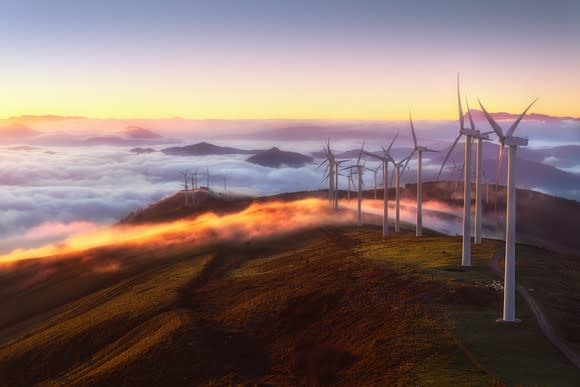 A row of wind turbines stretch across a mountain ridge as fog blankets the landscape behind it.