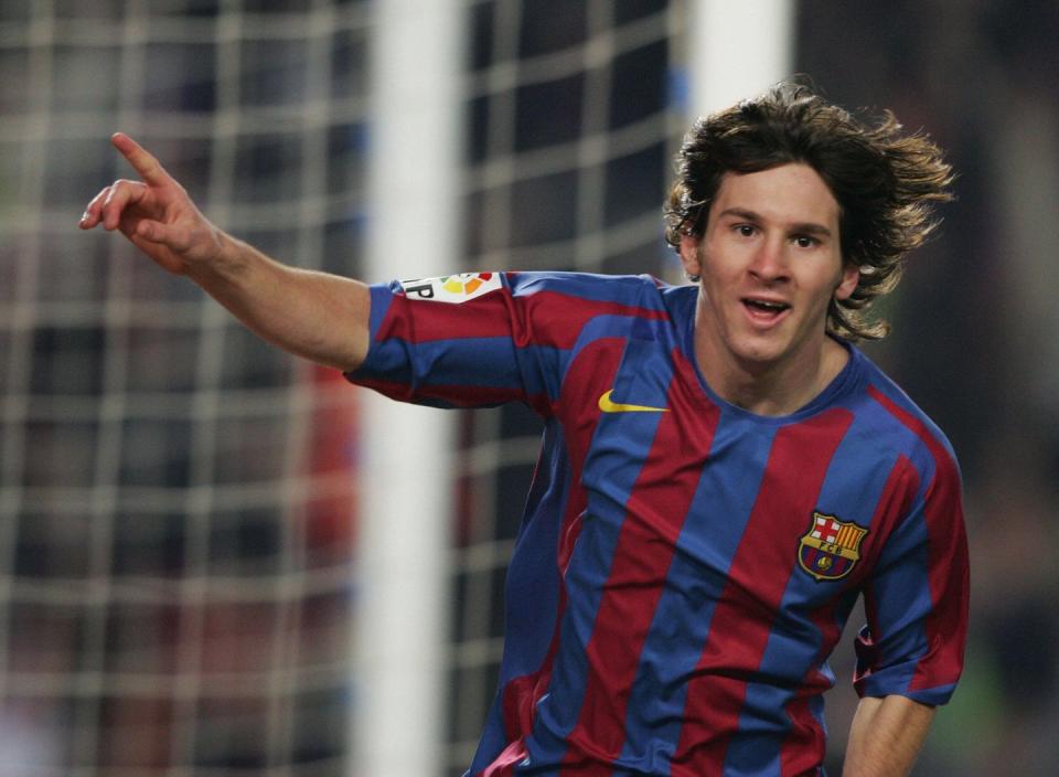 lionel messi looks at the camera with a smile on his face and right arm extended with his index finger pointed, he wears an fc barcelona jersey with blue and maroon stripes, behind him is a part of a blurry goal