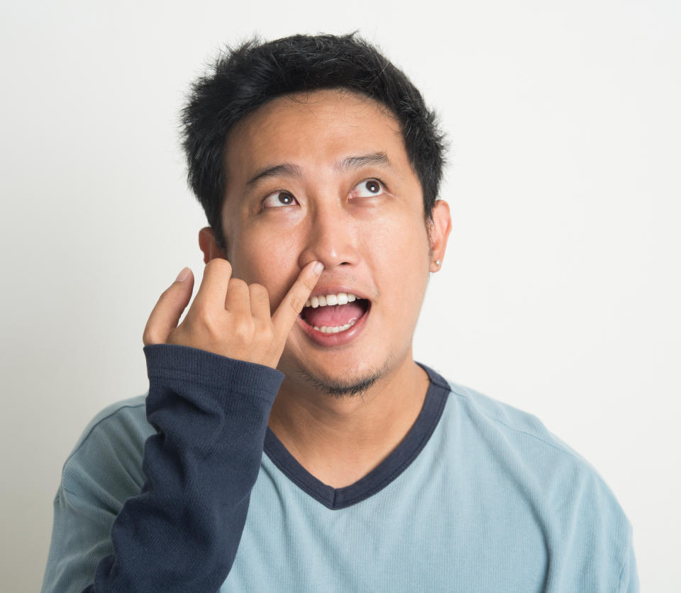 Disgusting Asian man picking nose with eyes looking up, on plain background