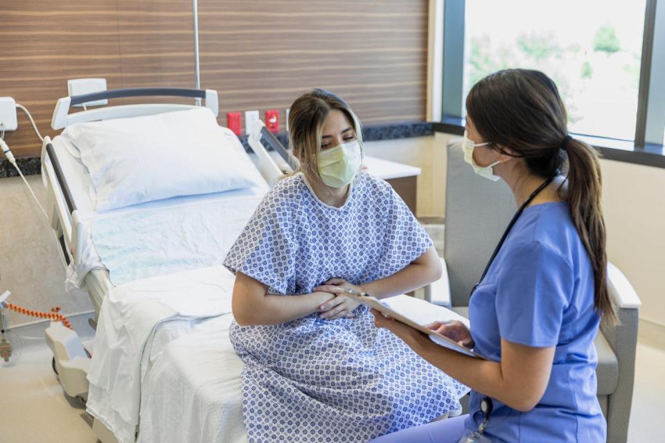 A woman in a medical mask and hospital gown sits on a bed holding her stomach as she speaks with a woman in blue scrubs.