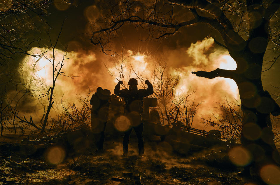 Ukrainian soldiers, standing in front of tall flames, fire artillery at Russian positions.