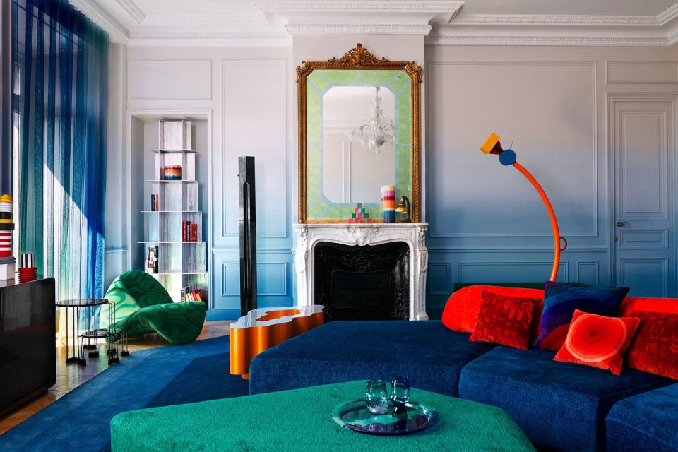 a very colorful living room with bright jewel blue wraparound sofa with large red throw pillows and green ottoman and modern and blue rug and transparent blue curtains and a white fireplace