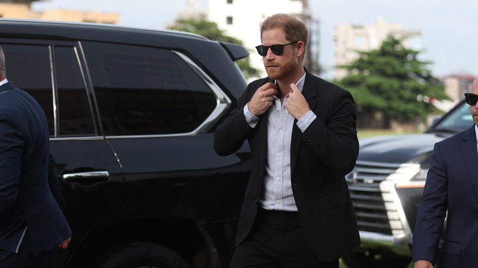 Prince Harry wearing sunglasses and fixing his tie next to a car.