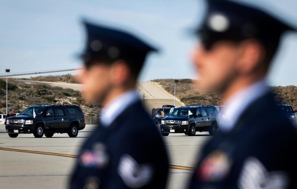 Two people in dark uniforms stand in the foreground while black SUVs travel in the background.