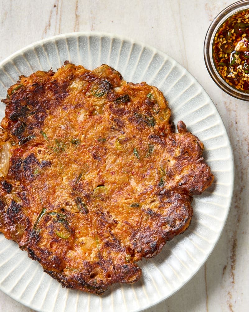 overhead shot of an uncut kimchi pancake on a white plate, with a bowl of dipping sauce to the side