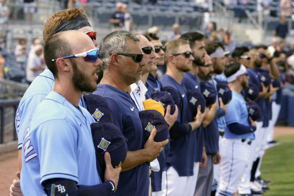 Tampa Bay Rays players along with manager Kevin Cash, third from left, stand at attention for the national anthem prior to a spring training baseball game against the Atlanta Braves Saturday March 19, 2022, in Port Charlotte, Fla. (AP Photo/Steve Helber)