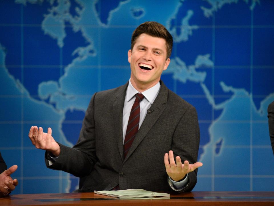 Colin Jost sitting at a desk and exclaiming while wearing a grey suit with a red tie on an episode of "Saturday Night Live"