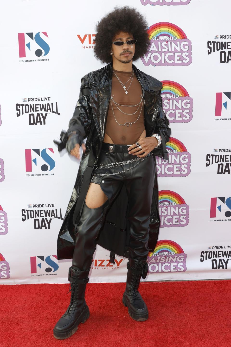 Keiynan Lonsdale poses on the red carpet in an all-black outfit with boots, leather pants, a mesh bodysuit, and long black coat, with sunglasses