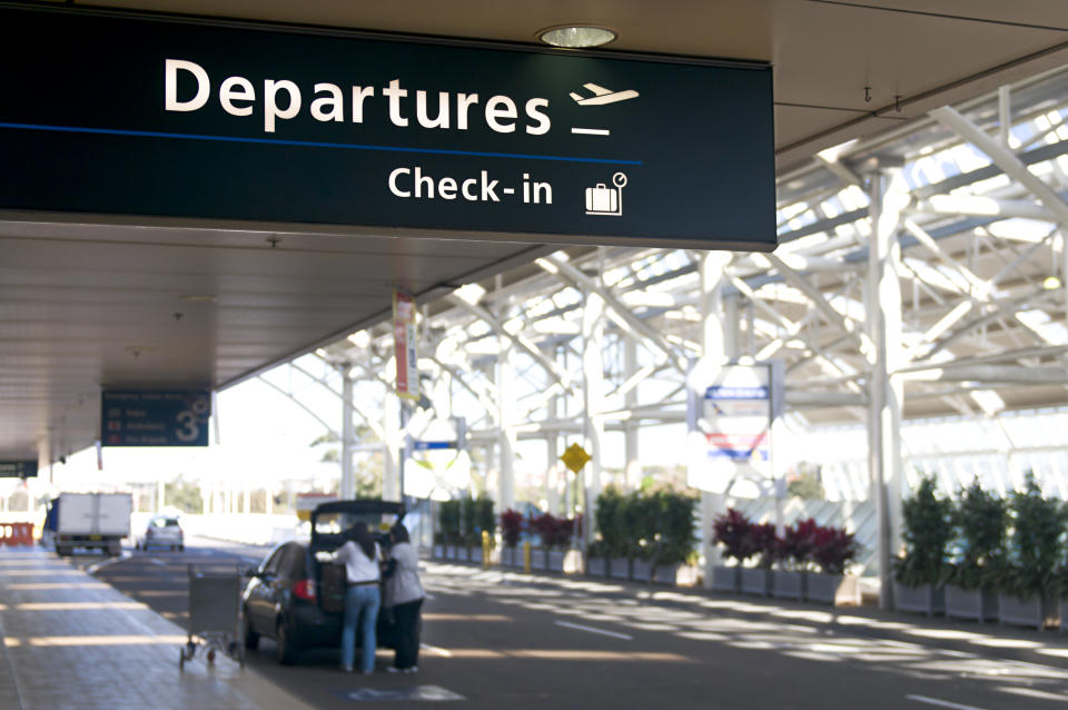 Sydney International Airport departures sign during the day with a car parked nearby and a suitcase being taken out. 