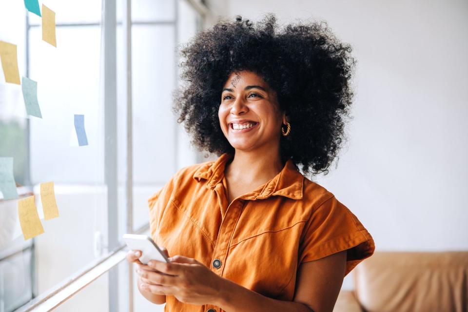 happy black businesswoman smiling while using a smartphone in a creative office cheerful female entrepreneur sending a text message while standing next to a glass wall with sticky notes