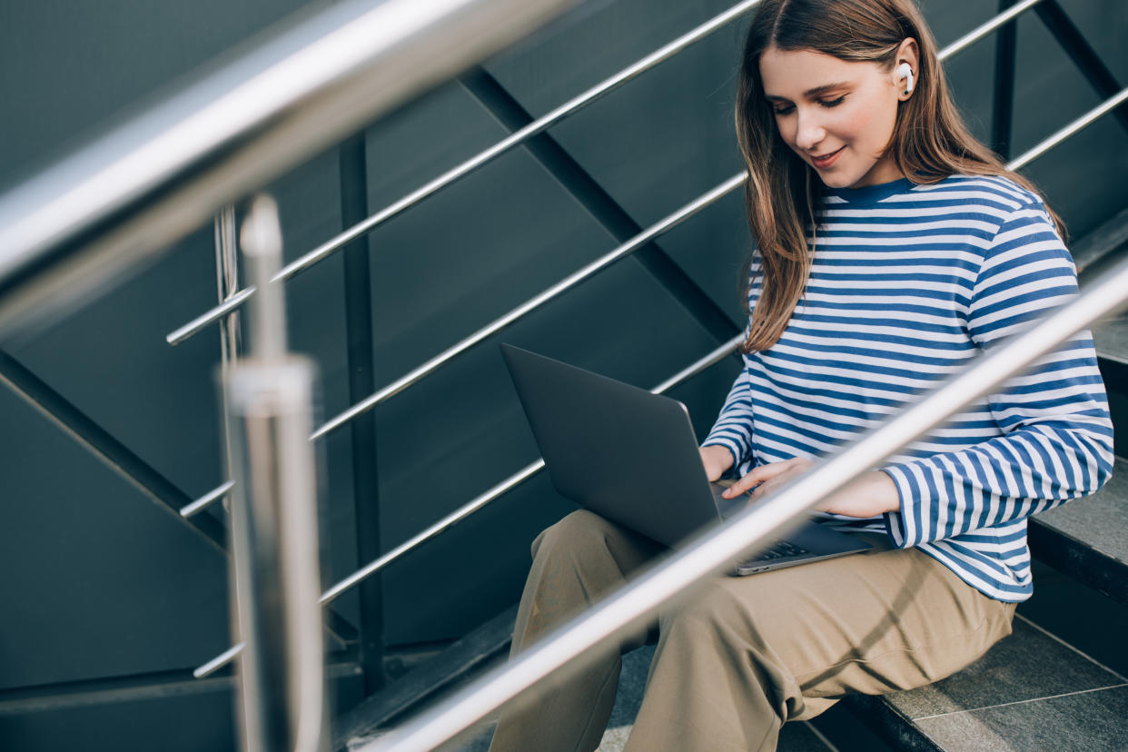 Millennial woman freelancer working online using her laptop, sitting on stairs.