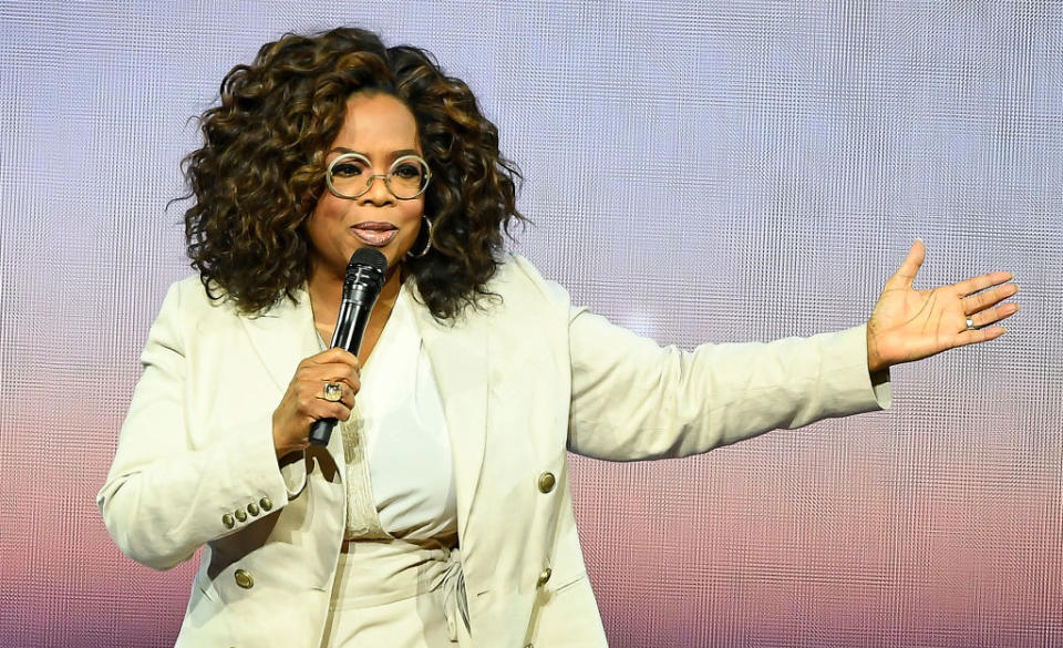 Oprah Winfrey speaking on stage, holding a microphone and gesturing with her other hand, wearing a light suit and glasses