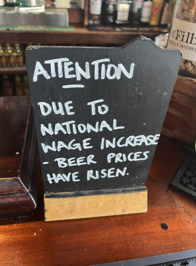 Sign reading "ATTENTION DUE TO WAGE INCREASE - BEER PRICES HAVE RISEN" on a bar counter