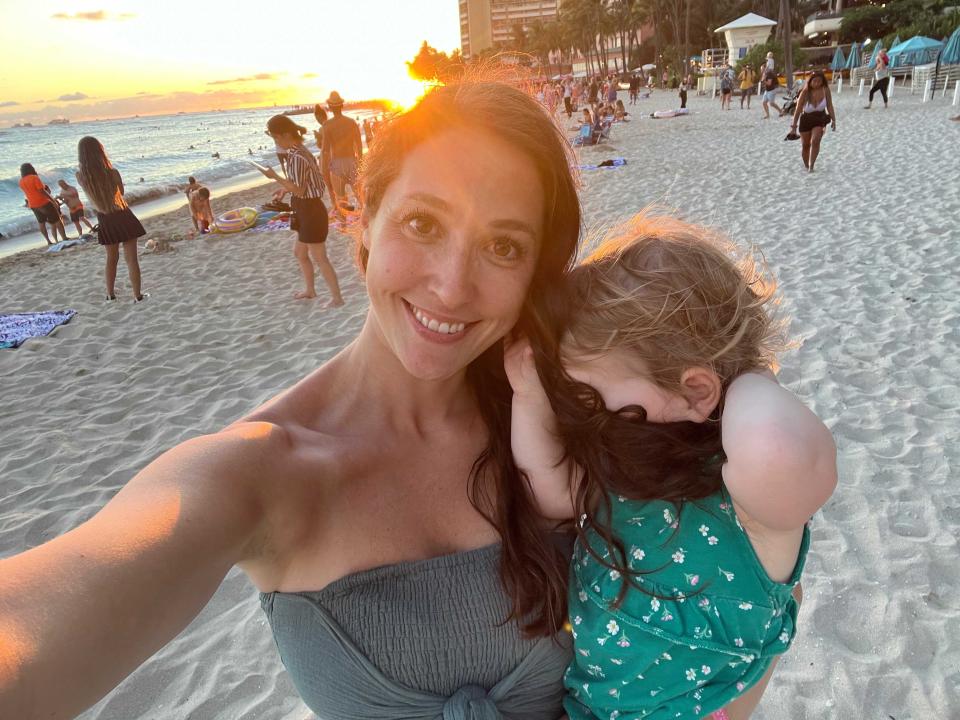 A woman taking a selfie at sunset while holding her child who is hiding behind her hair.