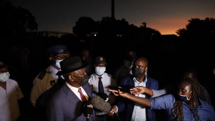 South Africa Ministry of Police, Bheki Cele, is surrounded by the press as he visits the crime scene after a botched cash-in-transit attack. The surroundings are dark.