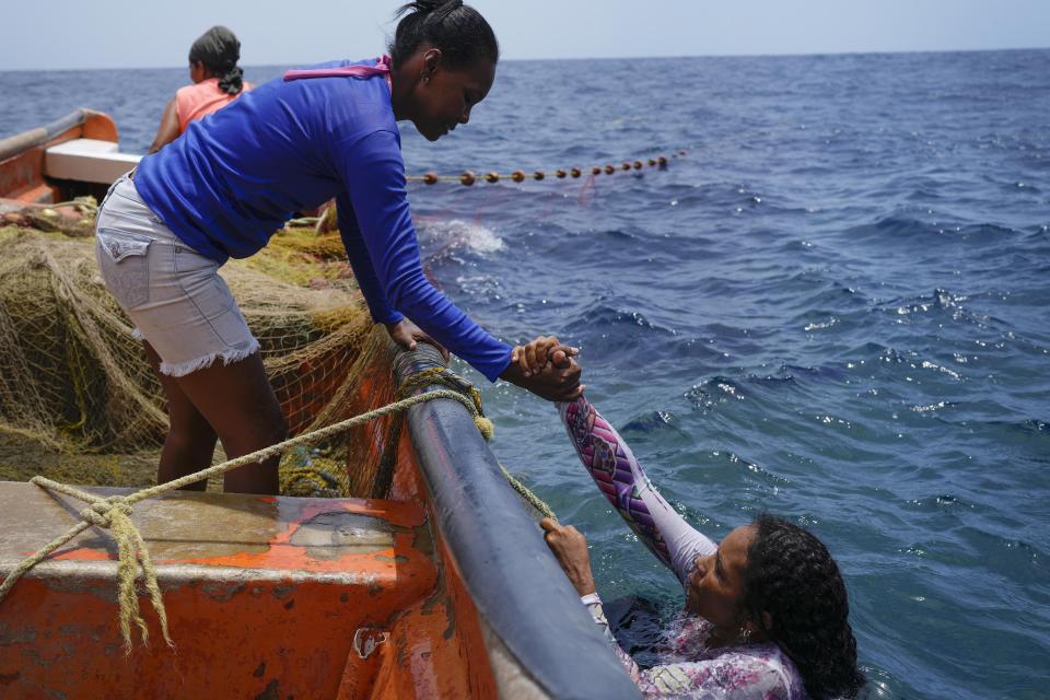 La pescadora Carolina Chávez sube a una lancha con la ayuda de una compañera frente a la costa de Tuja, Venezuela, el viernes 9 de junio de 2023. Chávez, quien empezó a pescar a los 11 años, estuvo a punto de perder su mano izquierda hace dos años cuando se enredó con una cuerda mientras ella y otros intentaban levantar una red pesada y sus botes chocaron. Cuando finalmente liberó su mano, la cuerda le había cortado la mitad de su dedo medio. (AP Foto/Matias Delacroix)