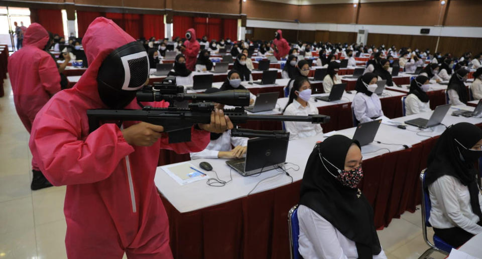 Exam participants are seated as people dressed as Squid Game guards hold up fake guns.