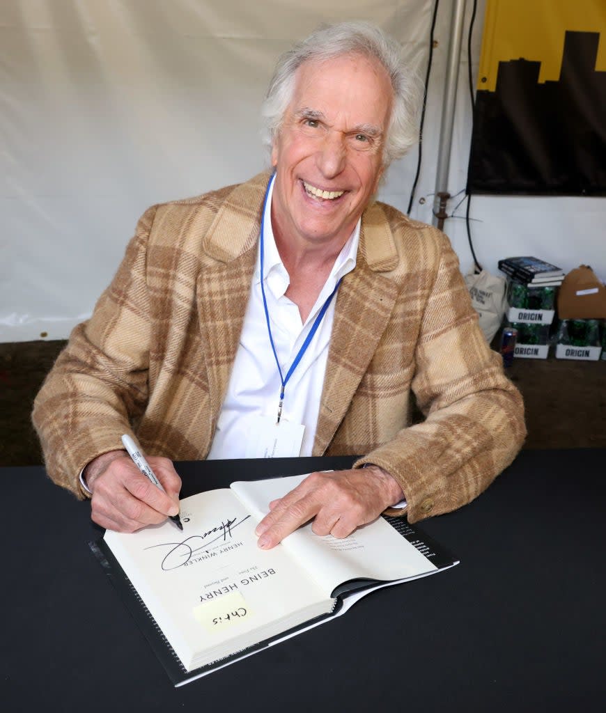 Henry Winkler smiles and signs a copy of his book at an event. He's wearing a plaid jacket, white shirt, and has a name badge around his neck