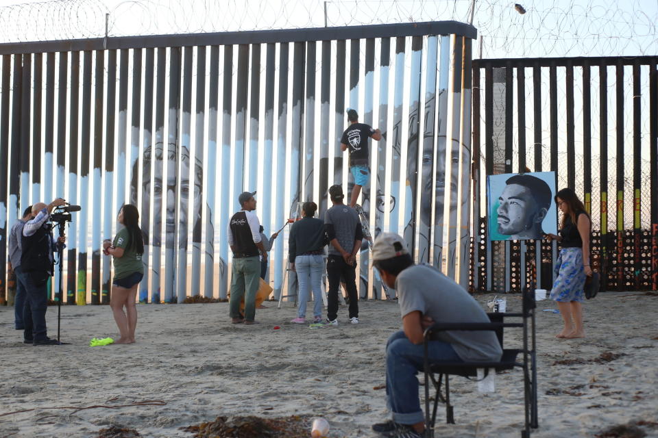 Artist Lizbeth De La Cruz Santana is videotaped during an interview, left, as volunteers help install her new mural on the Mexican side of a border wall that shows faces of people deported from the U.S. with barcodes that activate first-person narratives on visitors' phones, in Tijuana, Mexico, Friday, Aug. 9, 2019. De La Cruz Santana, 28, conceived the interactive mural as part of doctoral dissertation at University of California, Davis, in Spanish with a focus on literature and immigrant experiences.(AP Photo/Joebeth Terriquez)