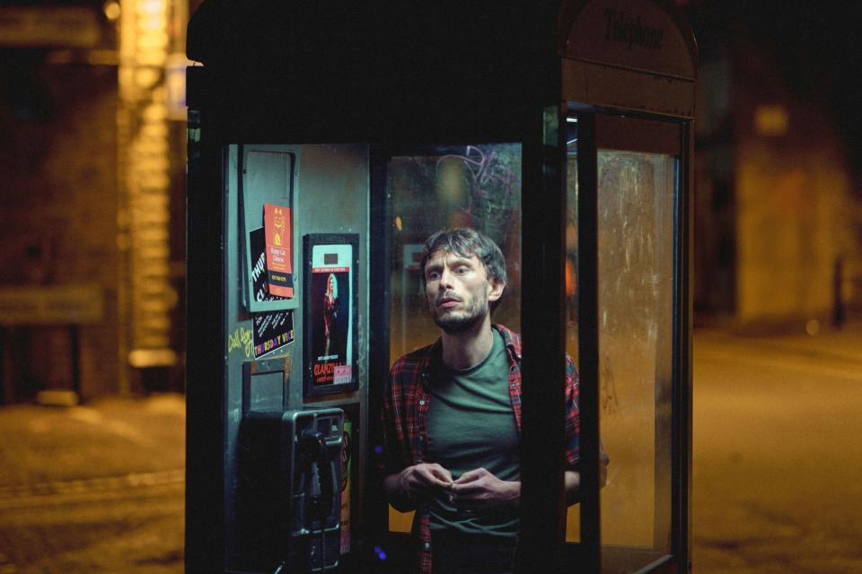 Richard Gadd in a checkered shirt inside phone booth at night, expression of contemplation
