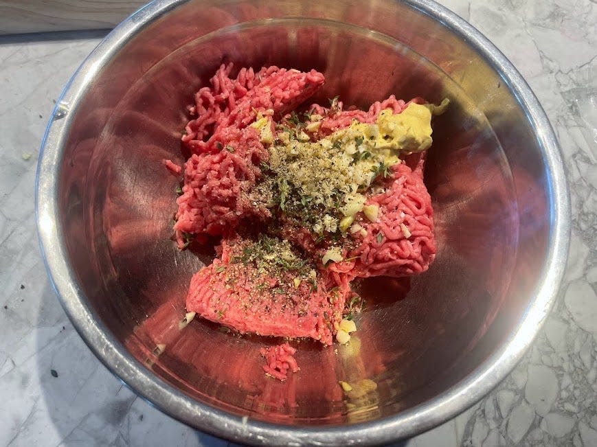 A metal bowl filled with ground beef, spices, and mustard. The spices sit on top of the meat and aren't mixed in yet