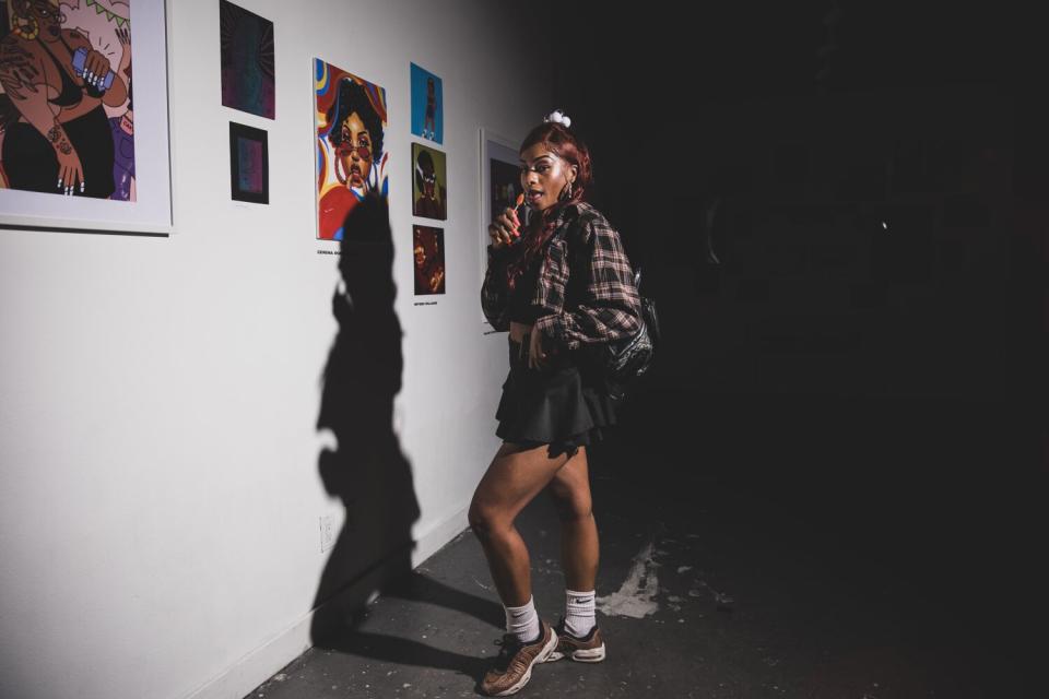 A person stands posing before a wall displaying art.