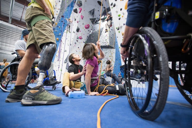 a gathering of climbers on the floor of a gym. One stands with a prosthesis. One sits in a wheelchair. A woman sits on the floor, belaying an invisible climber.