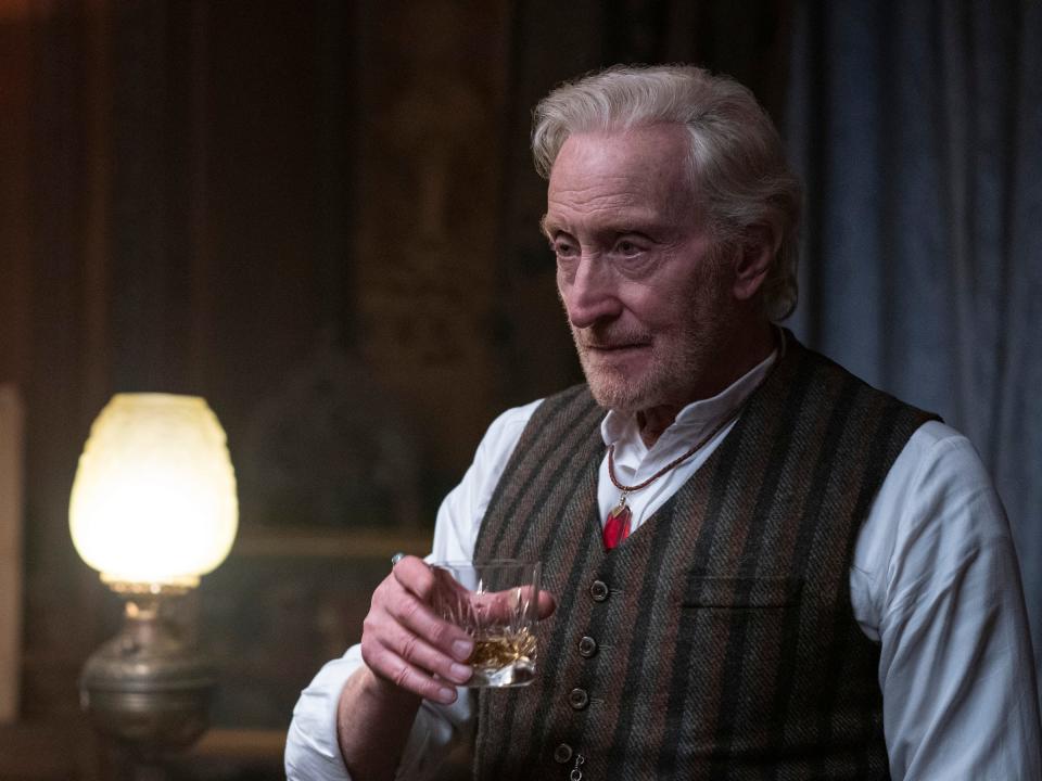 charles dance as roderick burgess in the sandman, wearing a button up shirt and vest and holding a small glass of whiskey